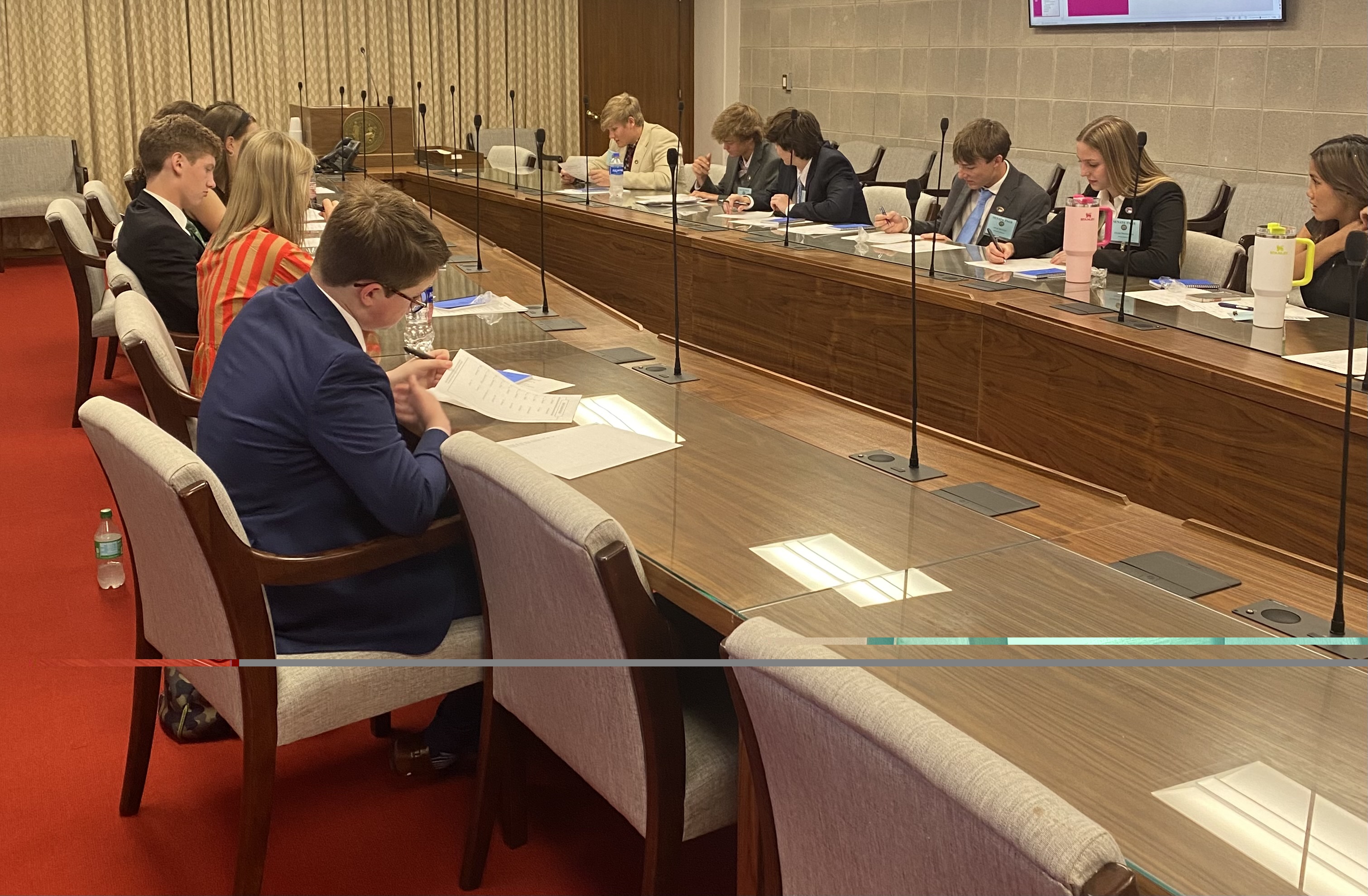 Pages seated at long table with microphones in committee room, looking at papers.